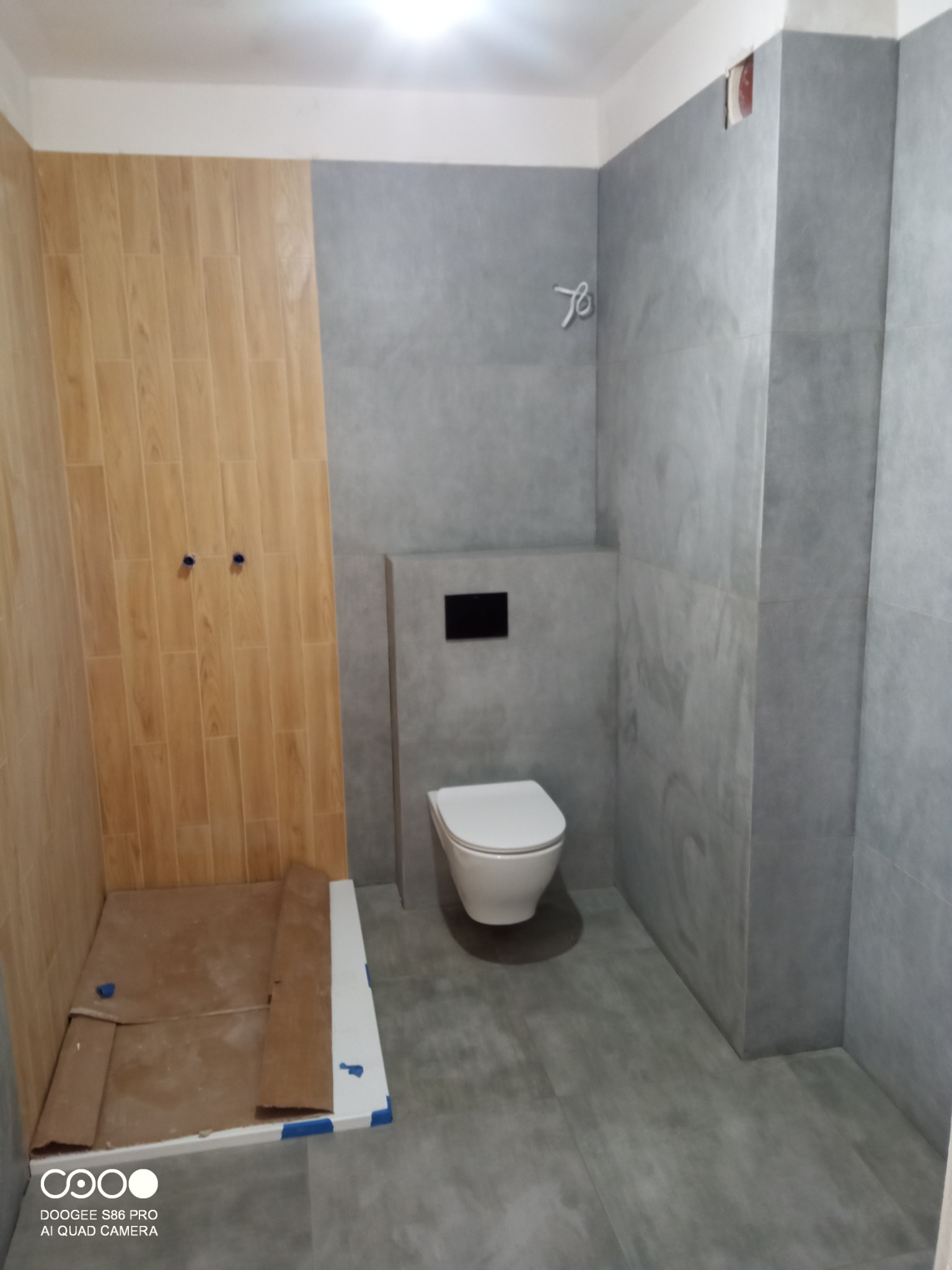 Wykończona łazienka z drewnopodobnymi i szarymi płytkami, białą toaletą. Widoczne przygotowane miejsce na prysznic. Minimalistyczny design.