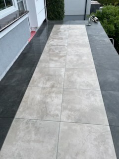 Wykończony taras z jasnoszarymi płytami na ciemnoszarym tle, widok z góry, częściowo widoczne rośliny w tle. Układ płytek wzdłuż długości tarasu.