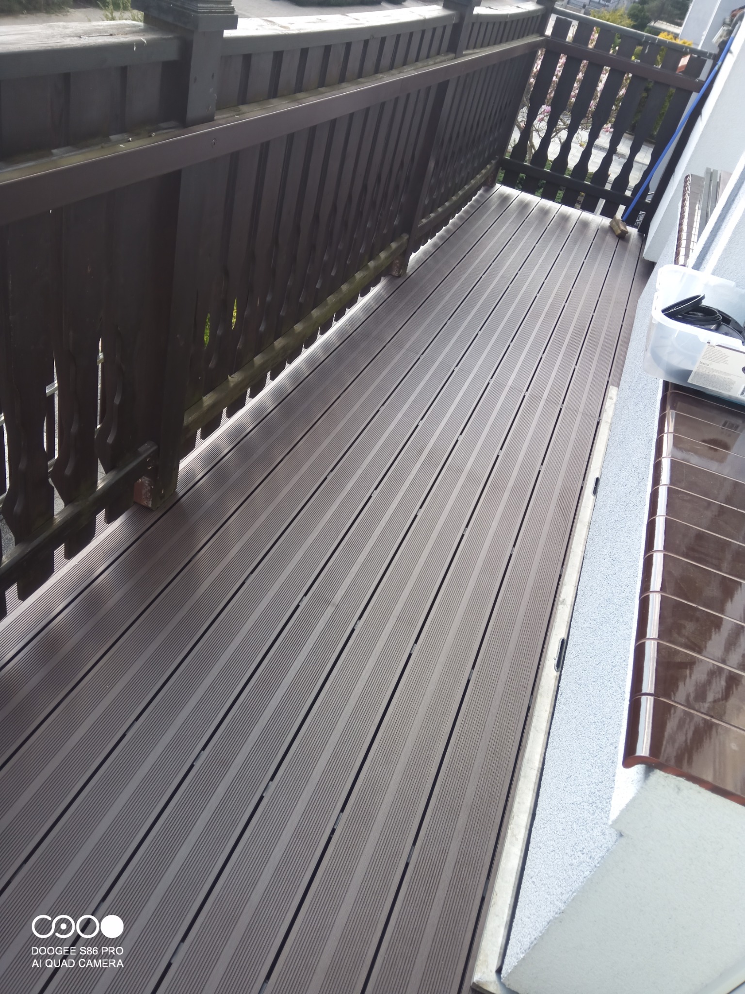 Nowoczesny balkon z brązową podłogą z desek kompozytowych, drewniane balustrady i jasne ściany. Widok na zewnątrz przez balustradę.