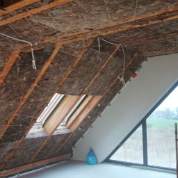 Brzozmal - Poddasze w trakcie ocieplania wełną mineralną, widoczne drewniane krokwie, siatka mocująca izolację, okno dachowe, betonowa wylewka na podłodze i drabina oparta o ścianę.