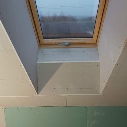 Brzozmal - Montaż okna dachowego w skośnej ścianie poddasza, widoczne płyty kartonowo-gipsowe w trakcie szpachlowania, zielona płyta GK wodoodporna w dolnej części ściany.