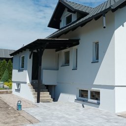 Brzozmal - Biała fasada domu jednorodzinnego z szarą dachówką i zadaszeniem nad wejściem, widok z poziomu kostki brukowej.