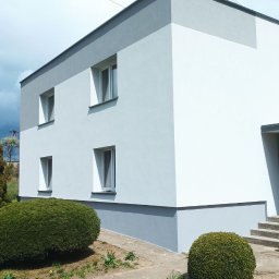 Brzozmal - Dwukondygnacyjny budynek po renowacji z szarą i białą elewacją, widoczne cztery okna, trawnik z krzewami i kwiatami, oraz fragment betonowego chodnika.