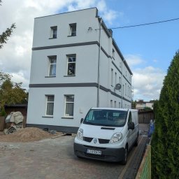 Brzozmal - Trzypiętrowy budynek po remoncie elewacji, pomalowany na szaro z ciemnoszarymi akcentami, zaparkowany biały samochód dostawczy, widoczny mieszalnik do betonu i pryzma piasku.