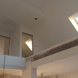 Brzozmal - Wnętrze domu w trakcie prac wykończeniowych: widoczne płyty kartonowo-gipsowe na ścianach i suficie, surowe otwory drzwiowe na antresoli oraz dwa okna dachowe wpuszczające jasne światło.