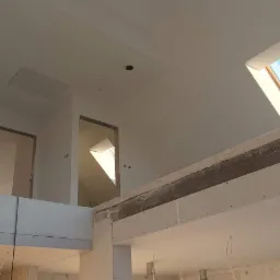 Wnętrze domu w trakcie prac wykończeniowych: widoczne płyty kartonowo-gipsowe na ścianach i suficie, surowe otwory drzwiowe na antresoli oraz dwa okna dachowe wpuszczające jasne światło.