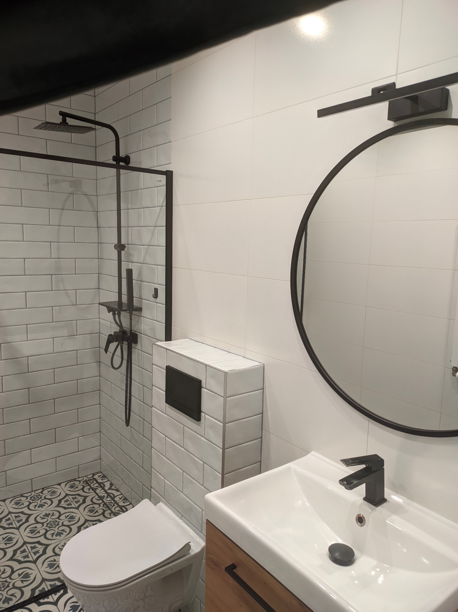 Nowoczesna łazienka z białymi płytkami imitującymi cegłę, czarną armaturą i okrągłym lustrem, wzorzysta podłoga dodaje charakteru.