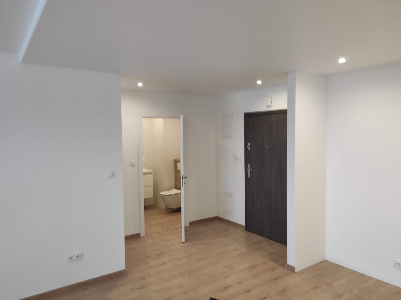Jasne, przestronne wnętrze po remoncie z drewnopodobną podłogą, widok na otwarte drzwi do łazienki z umywalką i toaletą, oraz ciemne drzwi wejściowe.