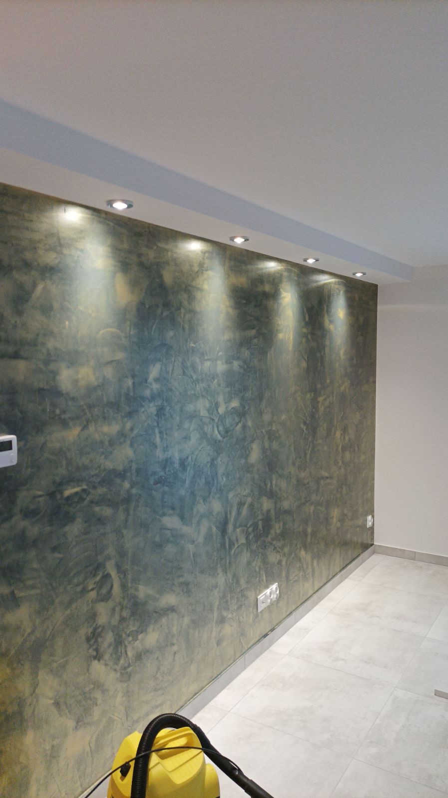 Ściana z dekoracyjną, ciemną gładzią w odcieniach szarości i złota, oświetlona punktowymi lampami sufitowymi, z widocznym odkurzaczem na pierwszym planie.