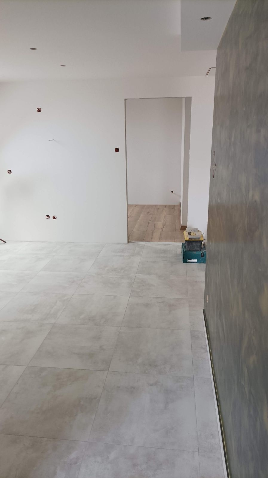 Wnętrze w trakcie prac wykończeniowych z szarymi płytkami podłogowymi, białą ścianą z widocznymi puszkami elektrycznymi i fragmentem ściany z efektem betonu; w tle widoczne narzędzia i podłoga...