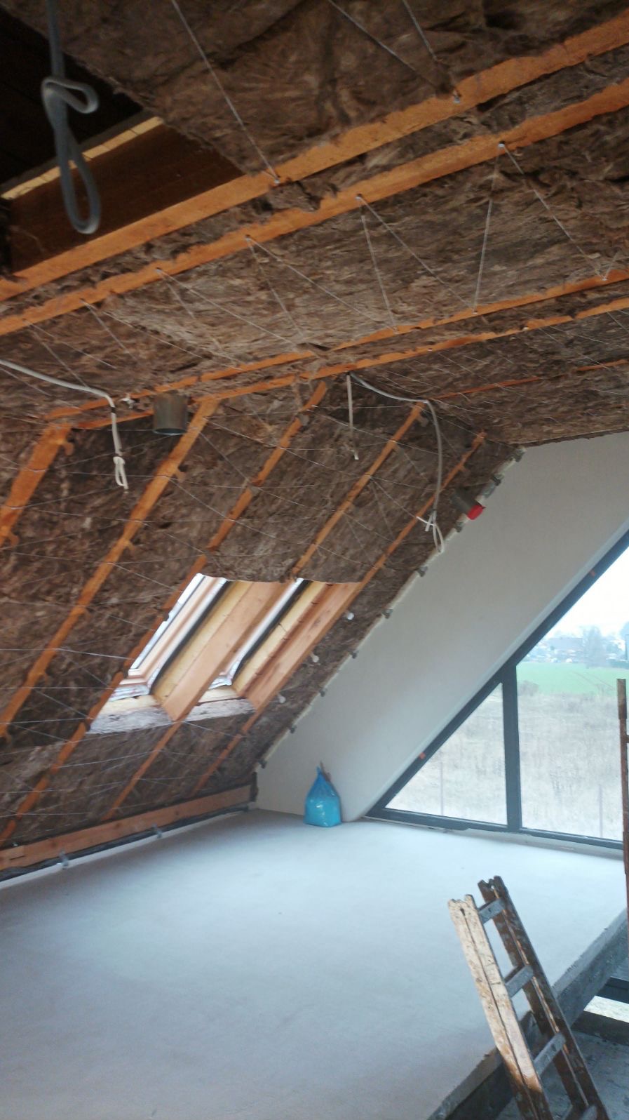 Poddasze w trakcie ocieplania wełną mineralną, widoczne drewniane krokwie, siatka mocująca izolację, okno dachowe, betonowa wylewka na podłodze i drabina oparta o ścianę.