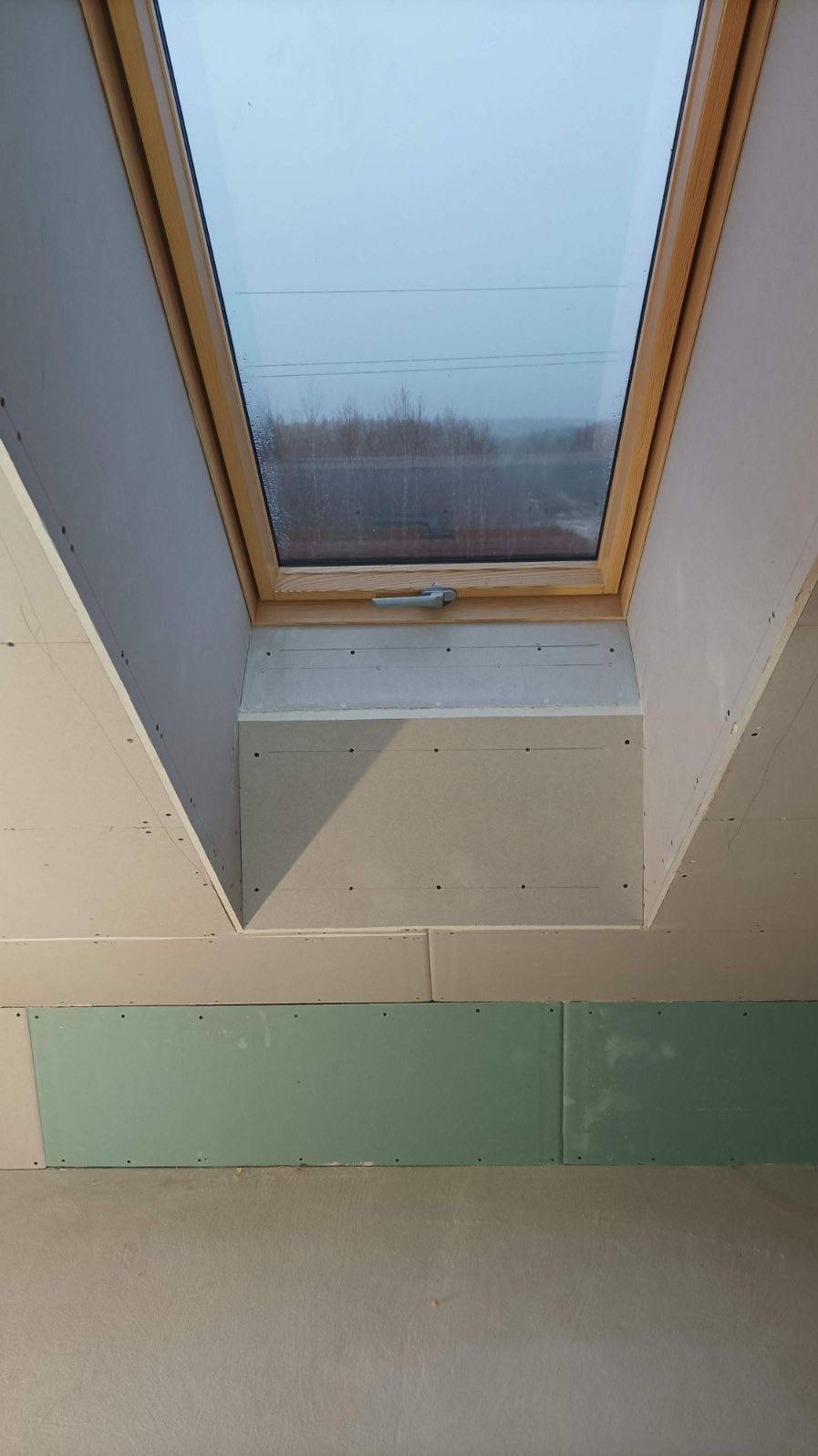 Montaż okna dachowego w skośnej ścianie poddasza, widoczne płyty kartonowo-gipsowe w trakcie szpachlowania, zielona płyta GK wodoodporna w dolnej części ściany.