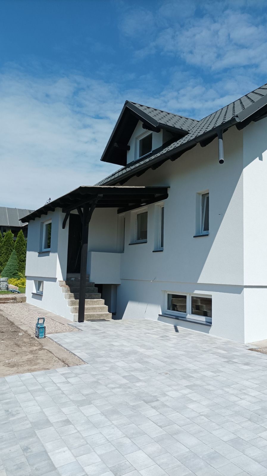 Biała fasada domu jednorodzinnego z szarą dachówką i zadaszeniem nad wejściem, widok z poziomu kostki brukowej.