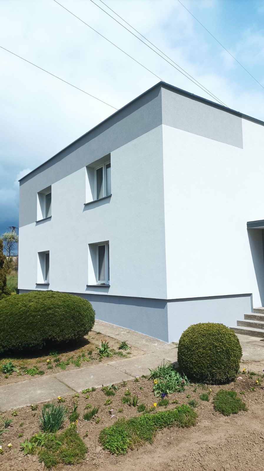 Dwukondygnacyjny budynek po renowacji z szarą i białą elewacją, widoczne cztery okna, trawnik z krzewami i kwiatami, oraz fragment betonowego chodnika.