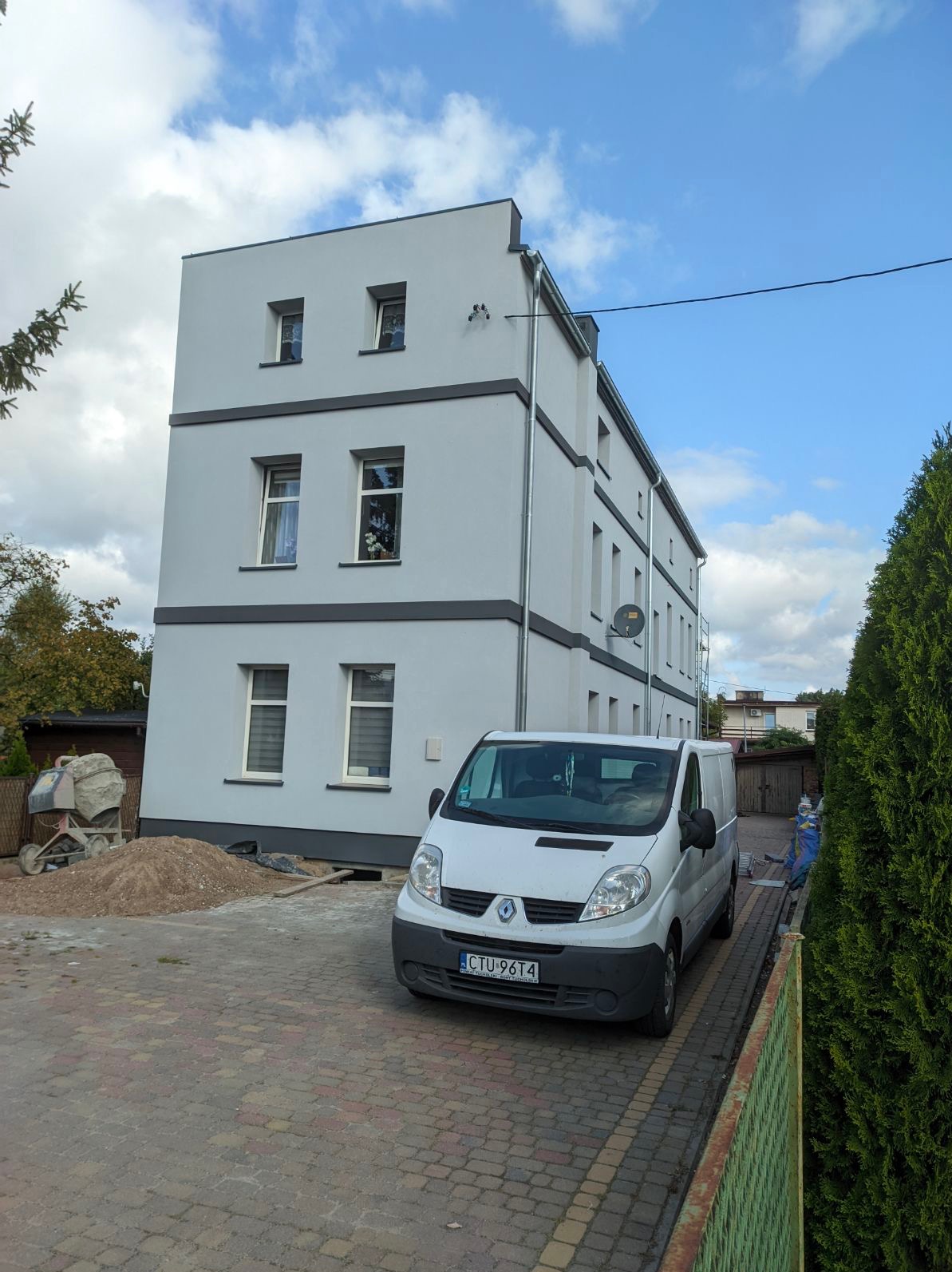 Trzypiętrowy budynek po remoncie elewacji, pomalowany na szaro z ciemnoszarymi akcentami, zaparkowany biały samochód dostawczy, widoczny mieszalnik do betonu i pryzma piasku.