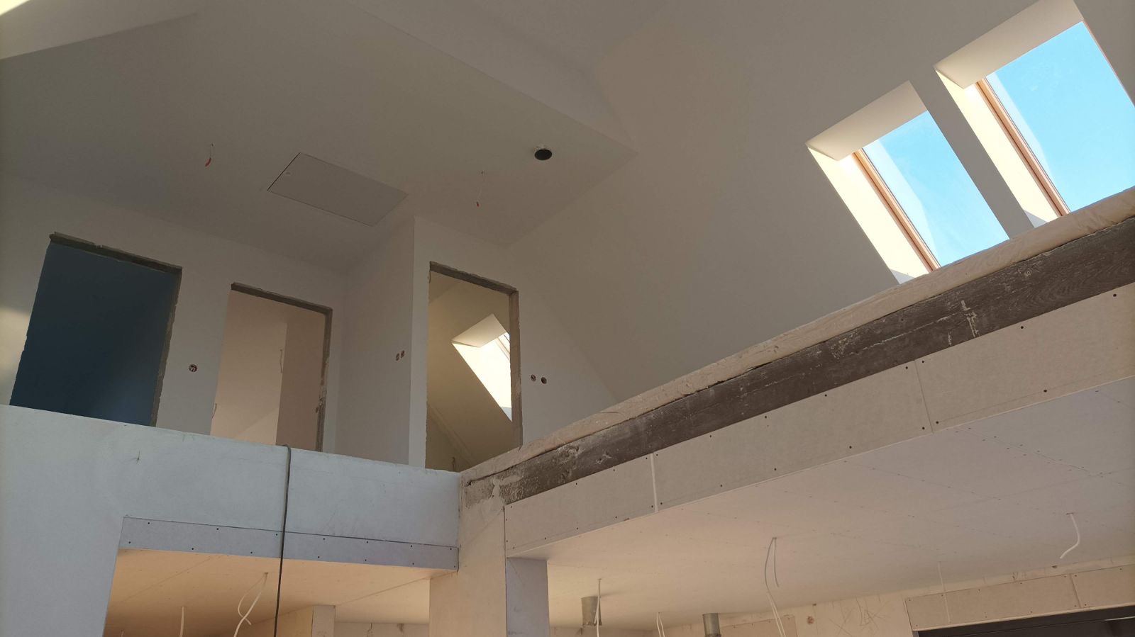 Wnętrze domu w trakcie prac wykończeniowych: widoczne płyty kartonowo-gipsowe na ścianach i suficie, surowe otwory drzwiowe na antresoli oraz dwa okna dachowe wpuszczające jasne światło.