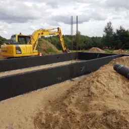 Żółta koparka Komatsu PL 130 obok wykopu pod fundamenty z czarną izolacją, widoczne zbrojenie i hałdy ziemi, w tle las i pochmurne niebo.