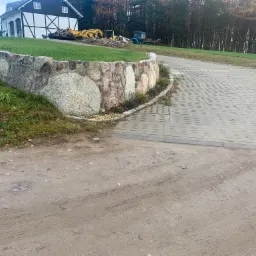 Ukończony kamienny murek oporowy przed posesją z kostką brukową, w tle maszyny budowlane i dom w stylu szachulcowym.