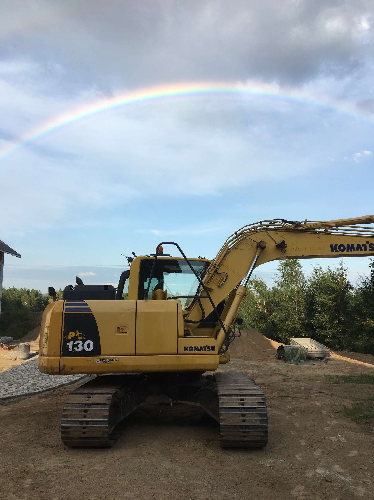 Żółta koparka Komatsu PC130 na placu budowy z widocznym fragmentem ułożonej kostki brukowej i tęczą na niebie.