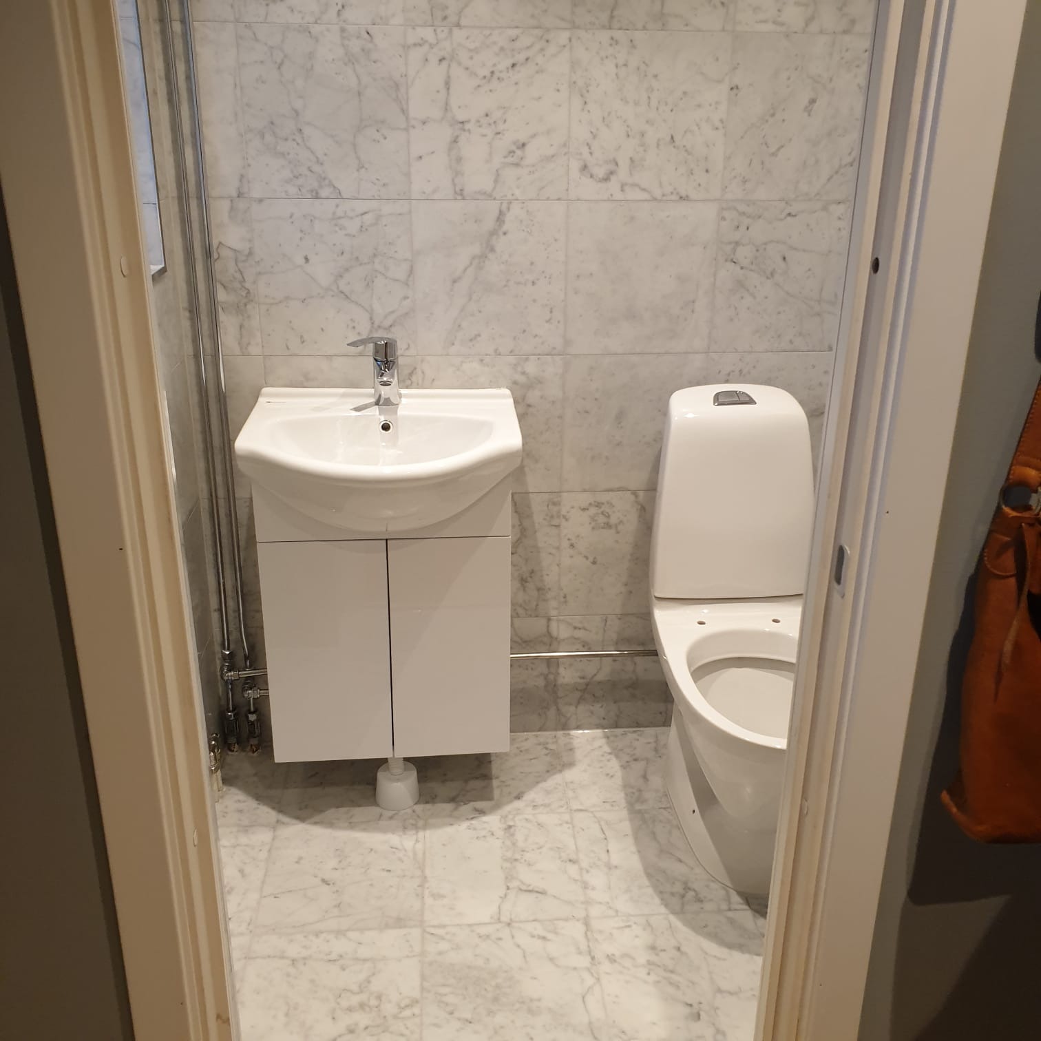 Wykończona łazienka z marmurowymi płytkami, białą umywalką z szafką i toaletą, widok zza framugi drzwi.