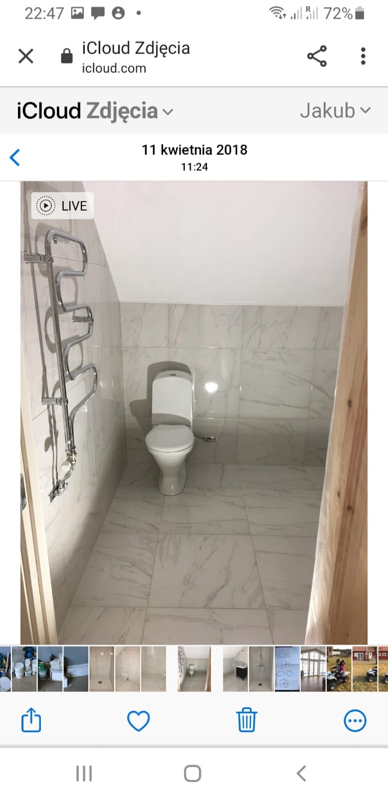 Wykończona łazienka z białymi marmurkowymi płytkami na ścianach i podłodze, z grzejnikiem drabinkowym i toaletą. Widoczny fragment drewnianej framugi drzwi.