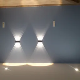 Dwa kinkiety LED zamontowane symetrycznie na gładkiej, pomalowanej na niebiesko ścianie, z widocznymi wiązkami światła tworzącymi stożki na ścianie i podłodze; w tle widoczna instalacja elektryczna.