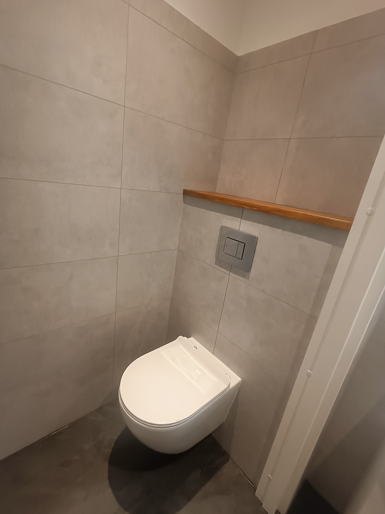 Wisząca toaleta w nowoczesnej łazience wykończonej szarymi, kwadratowymi płytkami ceramicznymi, z drewnianą półką nad spłuczką i widoczną częścią białej futryny drzwi.