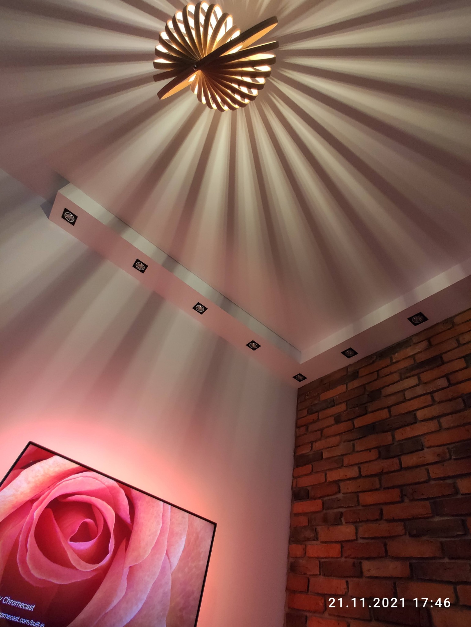 Drewniana lampa sufitowa z dekoracyjnymi cieniami, oświetlenie punktowe w listwie na suficie, fragment ściany z cegły i telewizor z wyświetlaną różą.