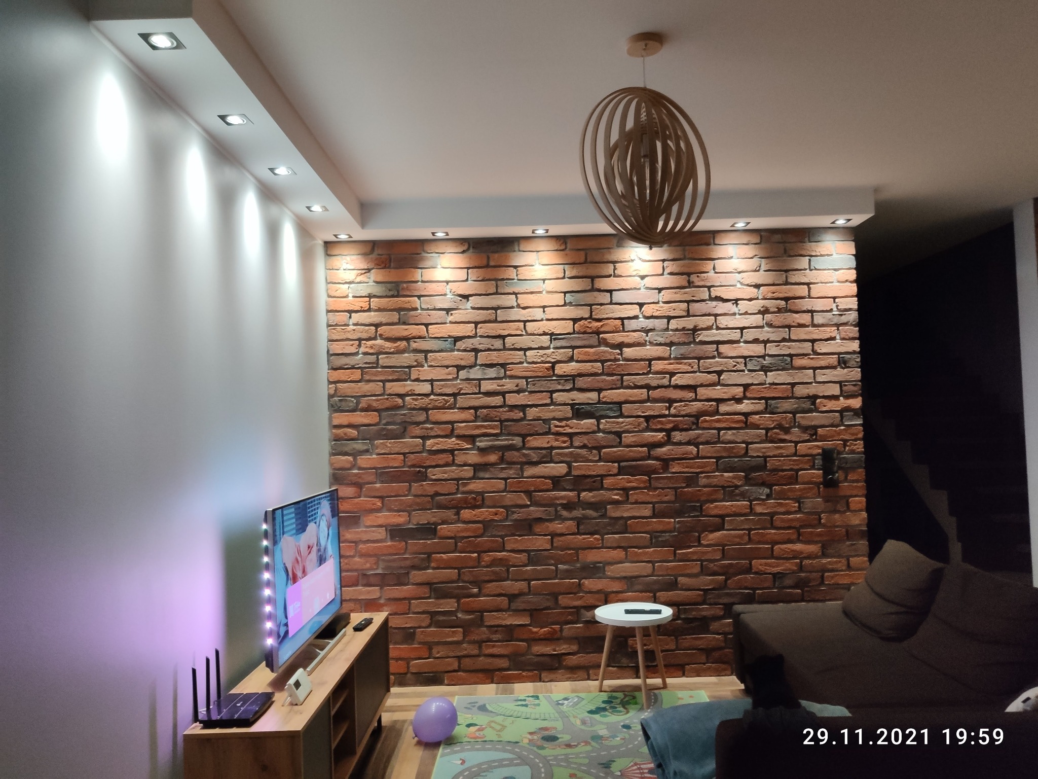 Salon z ceglaną ścianą, drewnianą lampą wiszącą, telewizorem podświetlonym LED i meblami w odcieniach brązu, oświetlony punktowo.