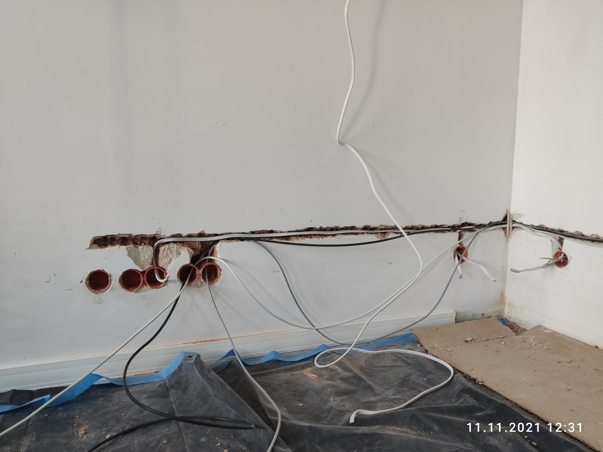 Ściana z wywierconymi otworami pod puszki elektryczne, widoczne przewody w trakcie instalacji, podłoga zabezpieczona folią malarską.