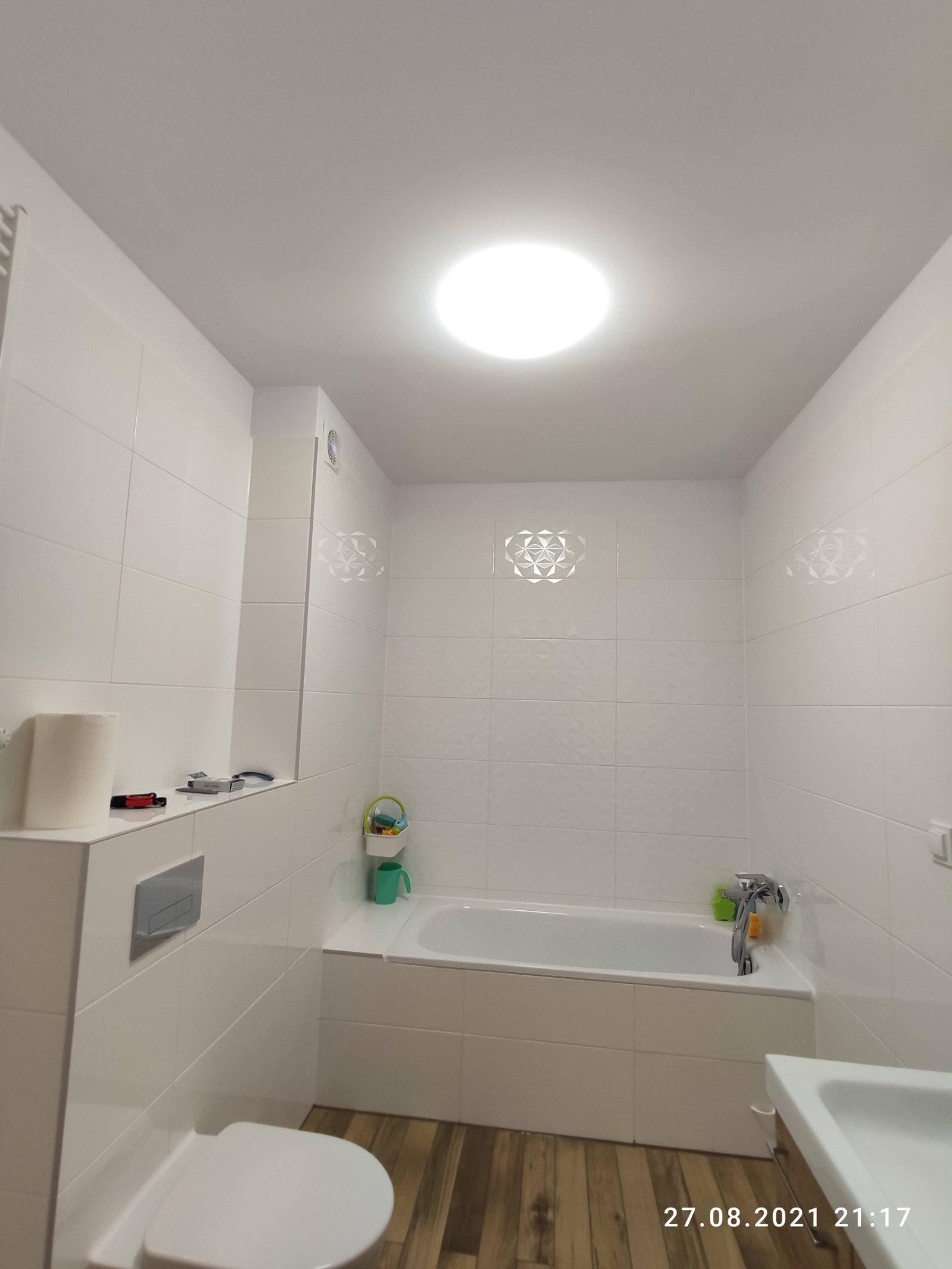 Nowoczesna, jasna łazienka z białymi płytkami, drewnopodobną podłogą i okrągłym oświetleniem sufitowym. Na ścianach dekoracyjne płytki z geometrycznym wzorem. Widoczna wanna, toaleta i umywalka.