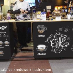 Be Honey Drukarnia - Zabudowa stoiska kawowego z tablicami kredowymi prezentującymi menu i rodzaje kaw, ozdobiona rysunkami filiżanek i ziaren kawy, widoczne ekspresy i akcesoria do parzenia kawy, w tle regały...