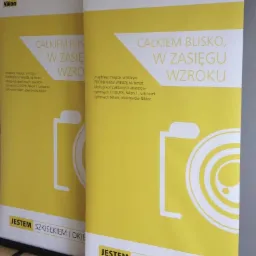 Dwa roll-upy reklamowe marki Nikon z żółto-białą grafiką, hasłem 'Całkiem blisko, w zasięgu wzroku' i uproszczonym rysunkiem aparatu fotograficznego.