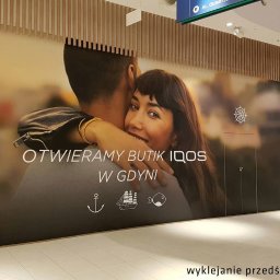 Be Honey Drukarnia - Wielkoformatowa grafika reklamowa na ścianie w galerii handlowej, przedstawiająca parę i informująca o otwarciu butiku IQOS w Gdyni, z motywami morskimi.