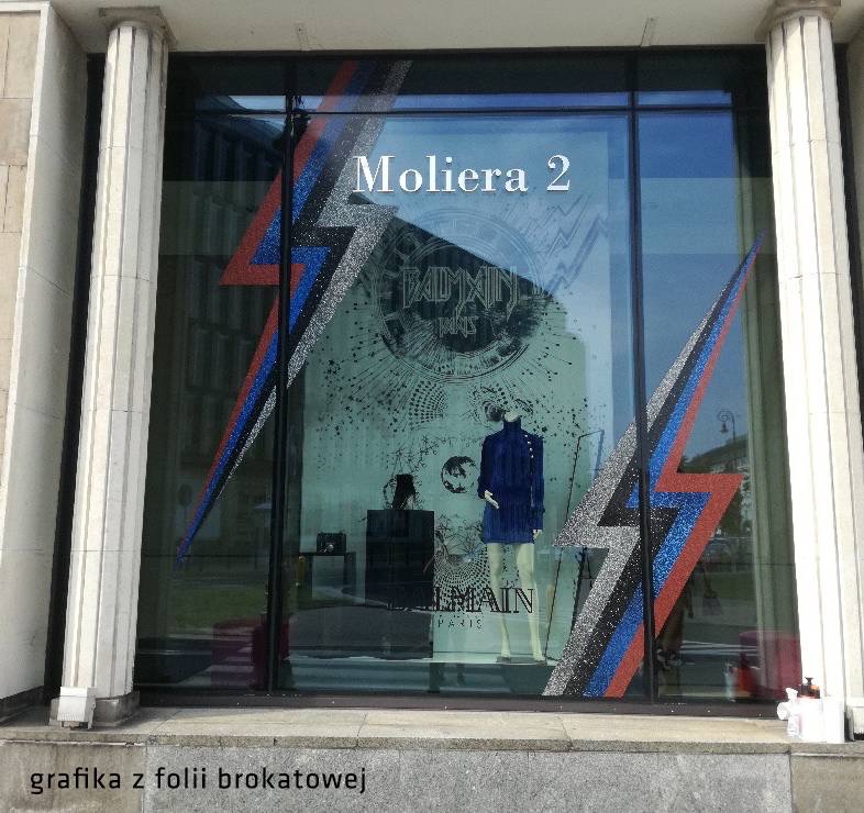 Witryna sklepu Balmain przy ulicy Moliera 2 ozdobiona grafiką z folii brokatowej w kształcie błyskawic w kolorach czerwonym, niebieskim i srebrnym, z logo marki i rysunkami planet w tle.
