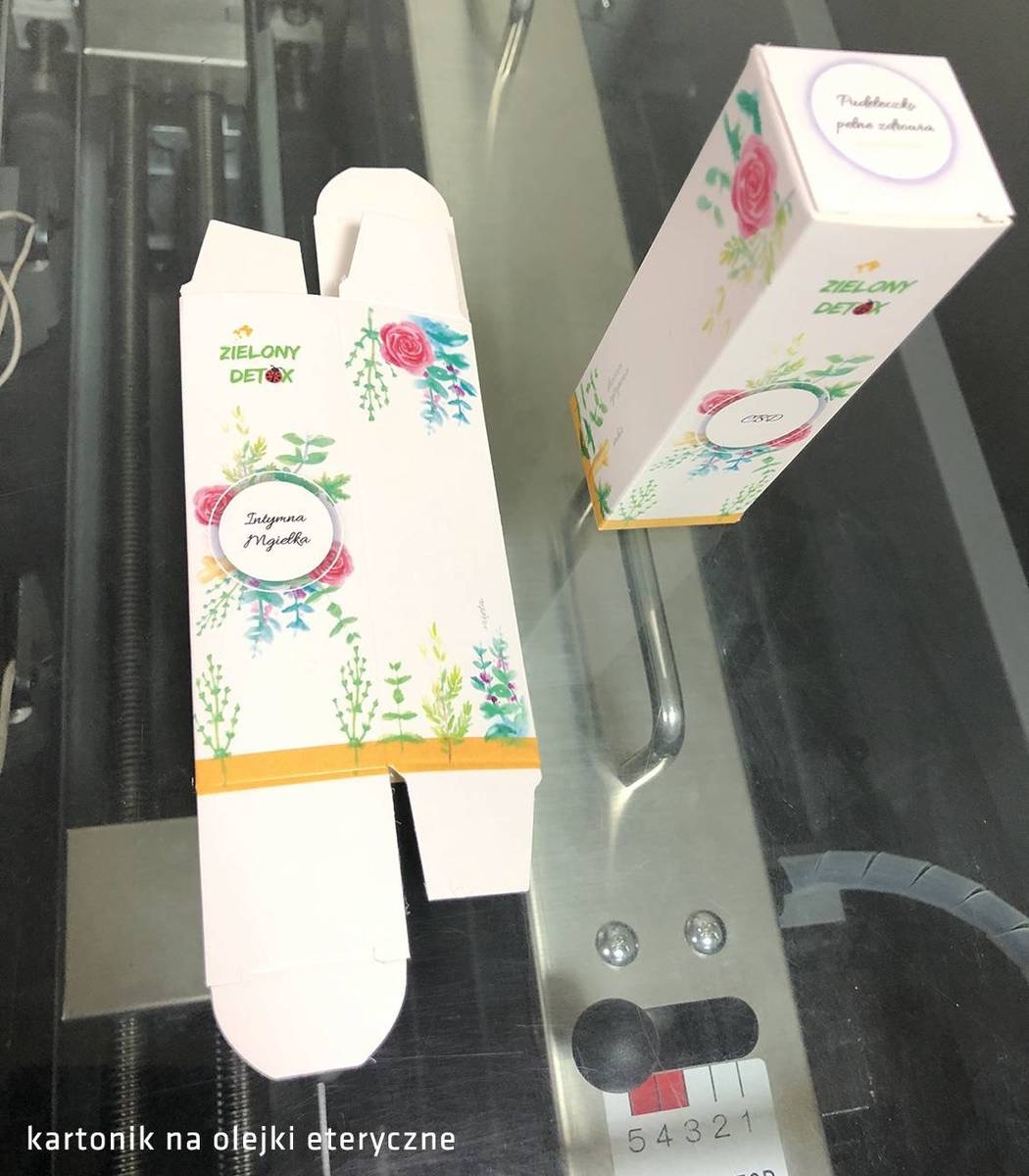 Dwa kartonowe pudełka z nadrukiem kwiatowym i logo 'ZIELONY DETOX', jedno złożone, drugie rozłożone, leżące na metalowej powierzchni maszyny.