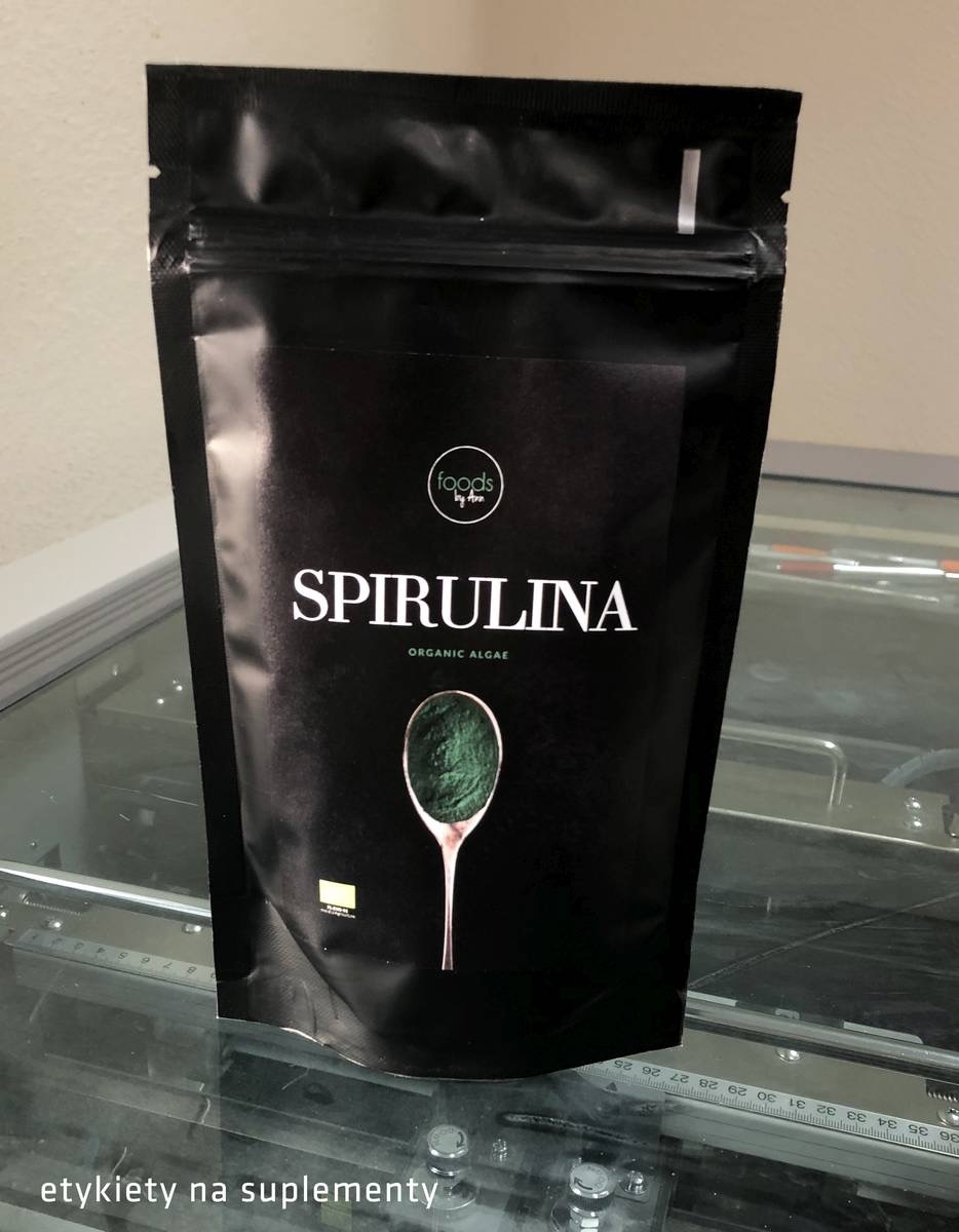 Czarna, błyszcząca torebka doypack z nadrukiem Spiruliny Organic Algae, prezentująca produkt na blacie z elementami maszyny.