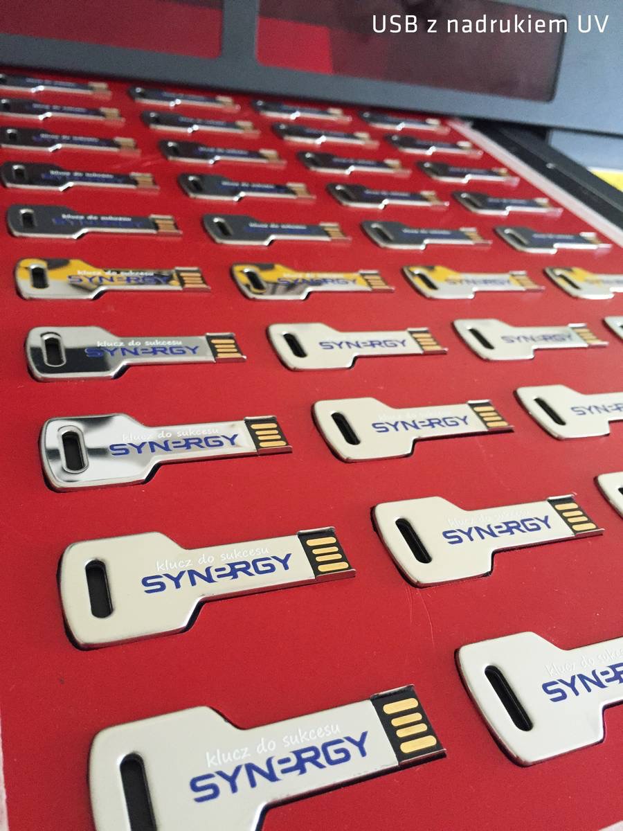 Partia pendrive'ów w kształcie klucza z nadrukowanym logo 'Synergy' na czerwonym podłożu, wykonanych techniką UV. Różne kolory i wzory nadruku.