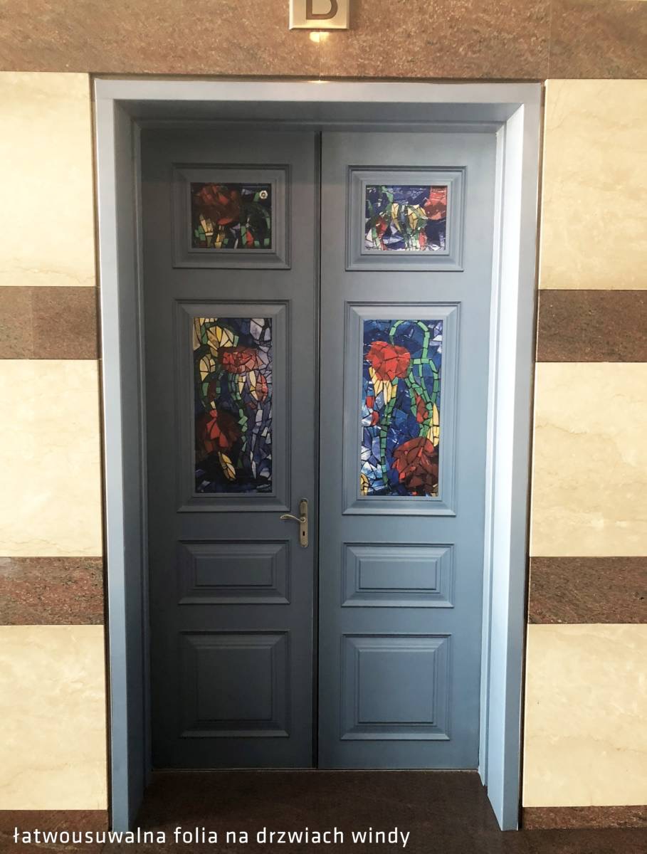 Szare dwuskrzydłowe drzwi windy z naklejonymi motywami kwiatowymi w stylu witrażu, umieszczonymi w kwadratowych i prostokątnych ramkach, na tle marmurowej ściany.