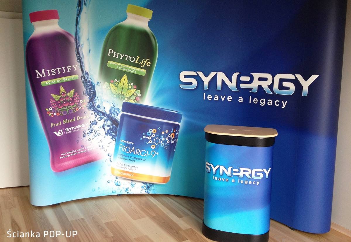 Zdjęcie przedstawia ściankę POP-UP z nadrukowanymi grafikami i logo marki Synergy oraz butelki napojów Mistify i PhytoLife oraz opakowanie ProArgi-9+ na drewnianej podłodze.