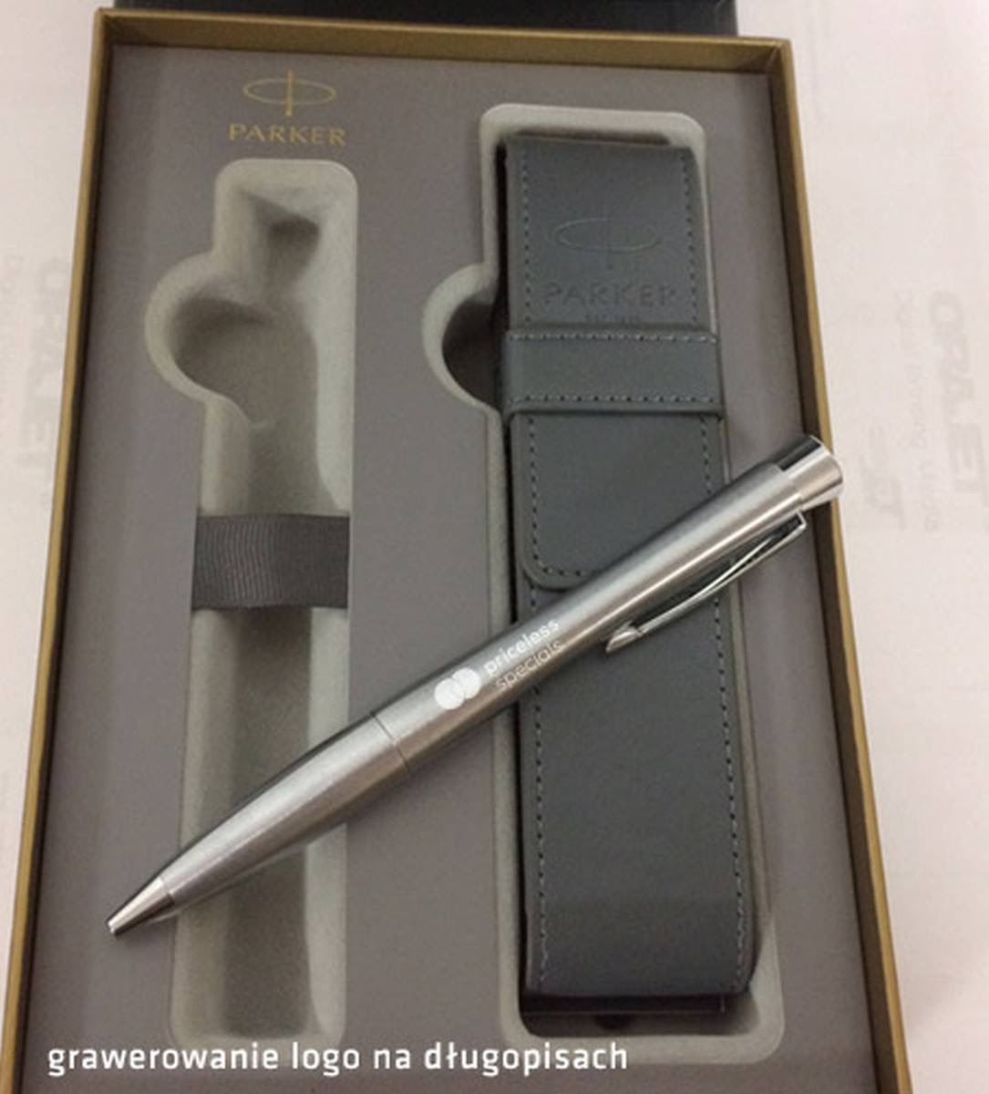 Zestaw upominkowy Parker: srebrny długopis z grawerowanym logo 'priceless specials' leżący na szarym skórzanym etui w pudełku.