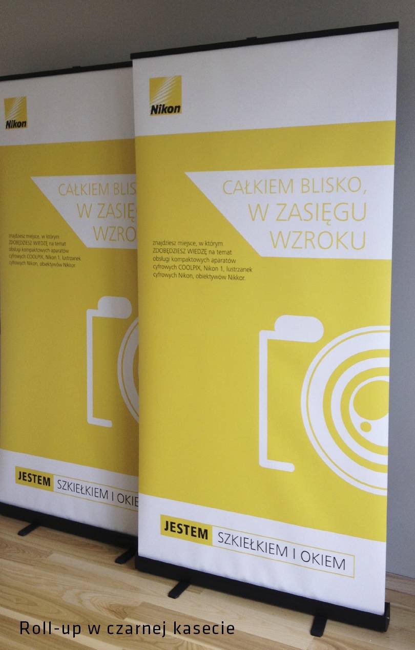 Dwa roll-upy reklamowe marki Nikon z żółto-białą grafiką, hasłem 'Całkiem blisko, w zasięgu wzroku' i uproszczonym rysunkiem aparatu fotograficznego.