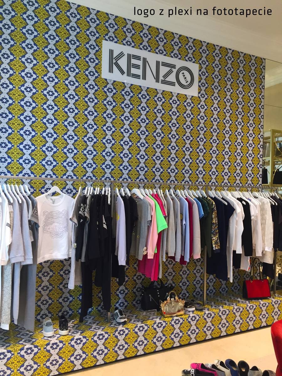 Wnętrze sklepu odzieżowego z fototapetą w żółto-niebieski wzór i podświetlanym logo marki na ścianie, w tle wieszaki z ubraniami i torebki na podeście.