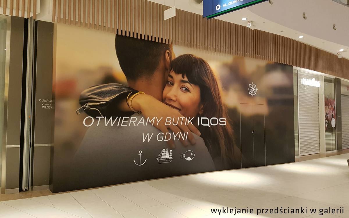 Wielkoformatowa grafika reklamowa na ścianie w galerii handlowej, przedstawiająca parę i informująca o otwarciu butiku IQOS w Gdyni, z motywami morskimi.