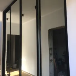 Czterodrzwiowa szafa wnękowa z lustrzanymi frontami i czarnymi ramami, odzwierciedlająca wnętrze w trakcie remontu z widocznym gruzem i narzędziami.