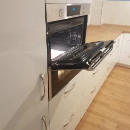 Biała zabudowa kuchenna z piekarnikiem z otwartymi drzwiczkami, widoczne blaty drewniane i parkiet na podłodze.