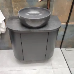 Czarna, okrągła umywalka nablatowa na szafce z zaokrąglonymi frontami, bateria w kolorze miedzi, lustro z widocznym odbiciem osoby, płytki imitujące rdzę na ścianie.