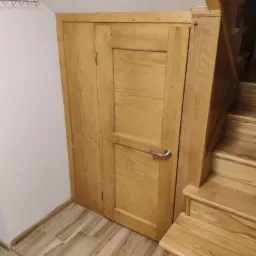 Drewniane schody z zabudową pod schodami, tworzącą schowek z drzwiami z prostym srebrnym uchwytem.