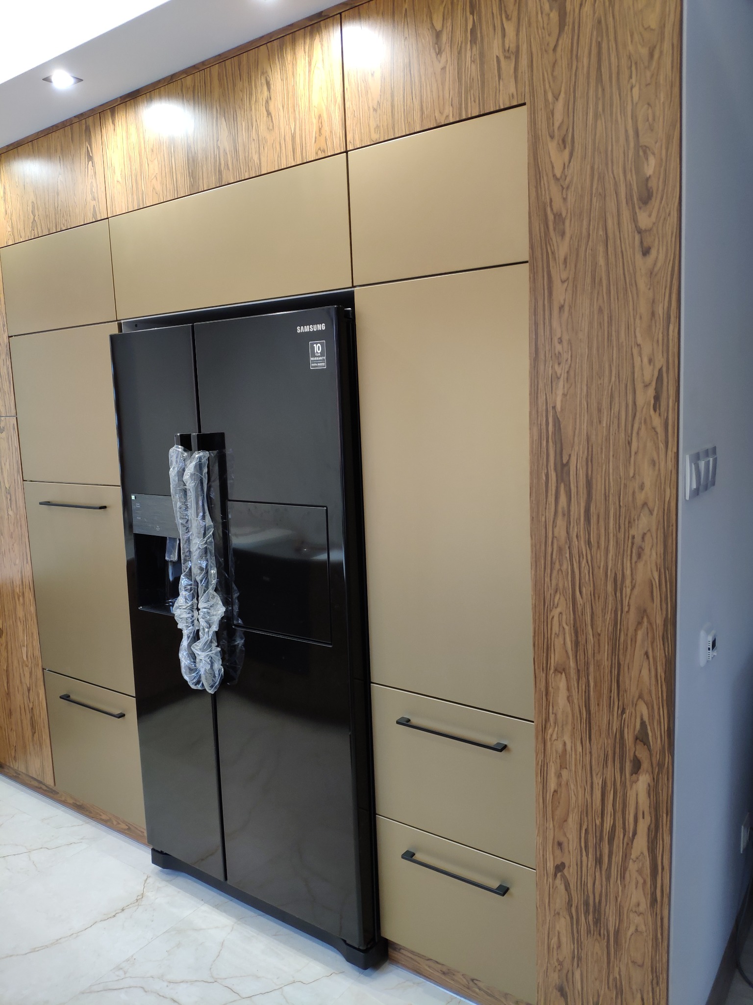 Czarna, dwudrzwiowa lodówka Samsung w zabudowie kuchennej z frontami w kolorze beżowym i drewnopodobnym, widoczne minimalistyczne uchwyty, marmurowa podłoga.