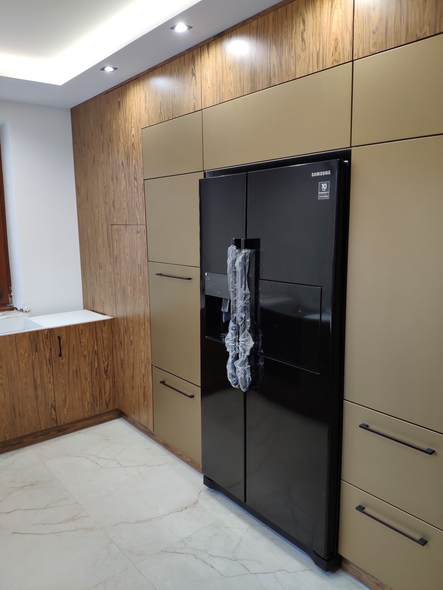 Nowoczesna zabudowa kuchenna w Spytkowicach z frontami w odcieniach drewna i beżu, z czarną lodówką typu side-by-side z folią ochronną na uchwytach.